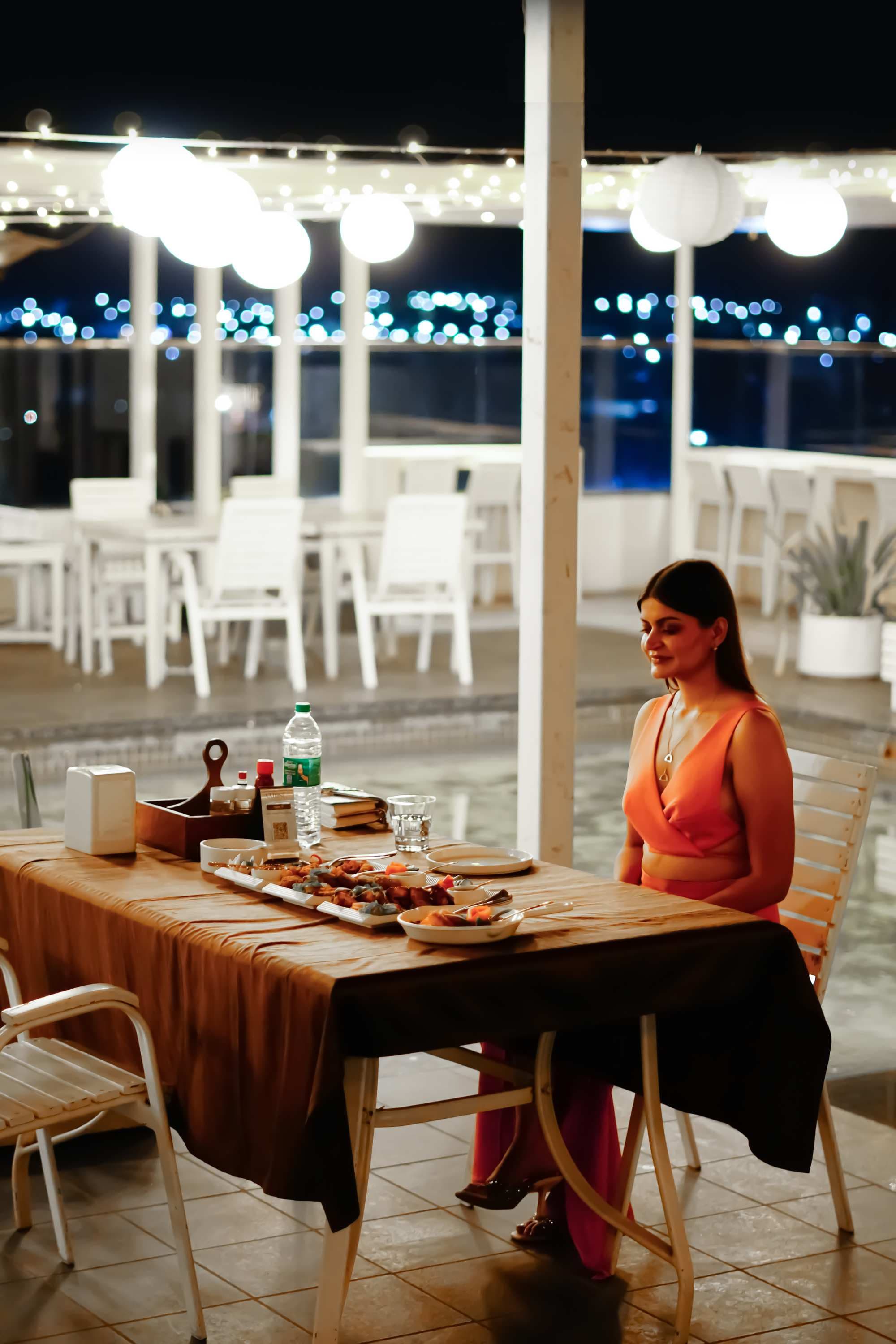 A woman at a poolside table at night, lights strung overhead