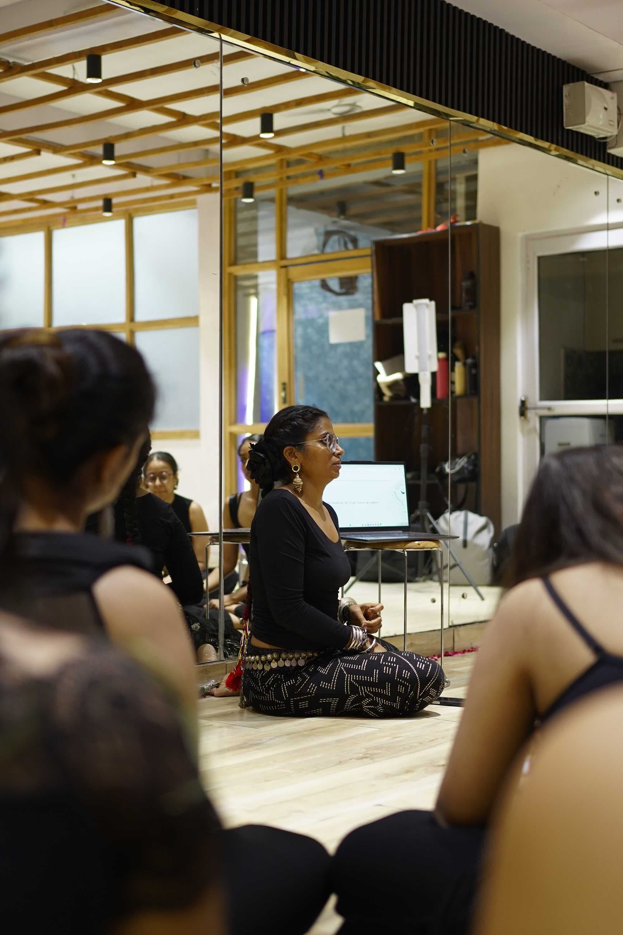 The instructor talking to the class, seated on the studio floor